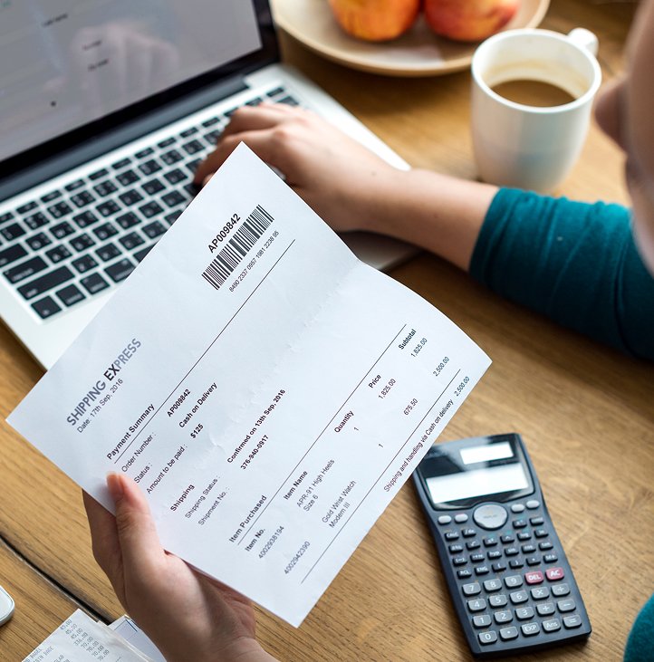 Person holding a shipping receipt, working on a laptop with a calculator and coffee on the desk.