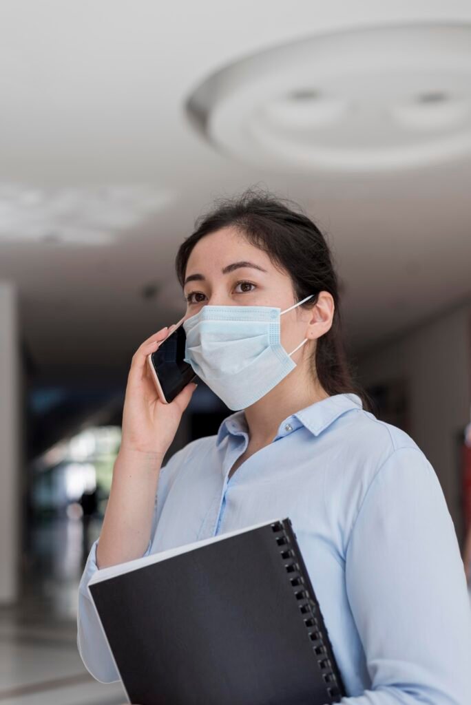 Professional woman in a mask, holding a notebook, speaking on the phone in an office setting.